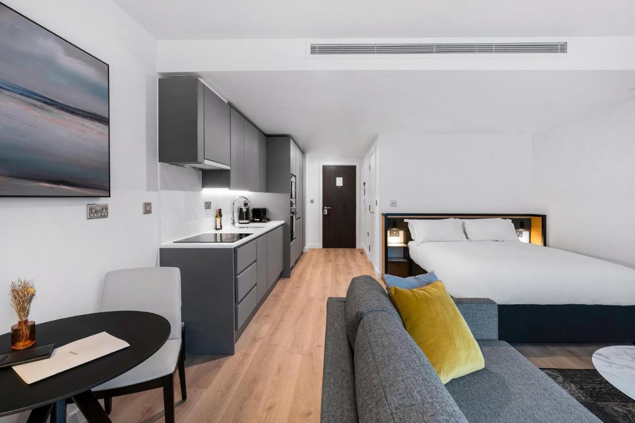 Citysuites 2 Aparthotel Manchester Kültér fotó The photo shows a modern and stylish studio apartment. It features a kitchenette with dark cabinetry and a small dining table with two chairs. There's a cozy sofa with colorful cushions, positioned next to the table. In the background, a large bed wi