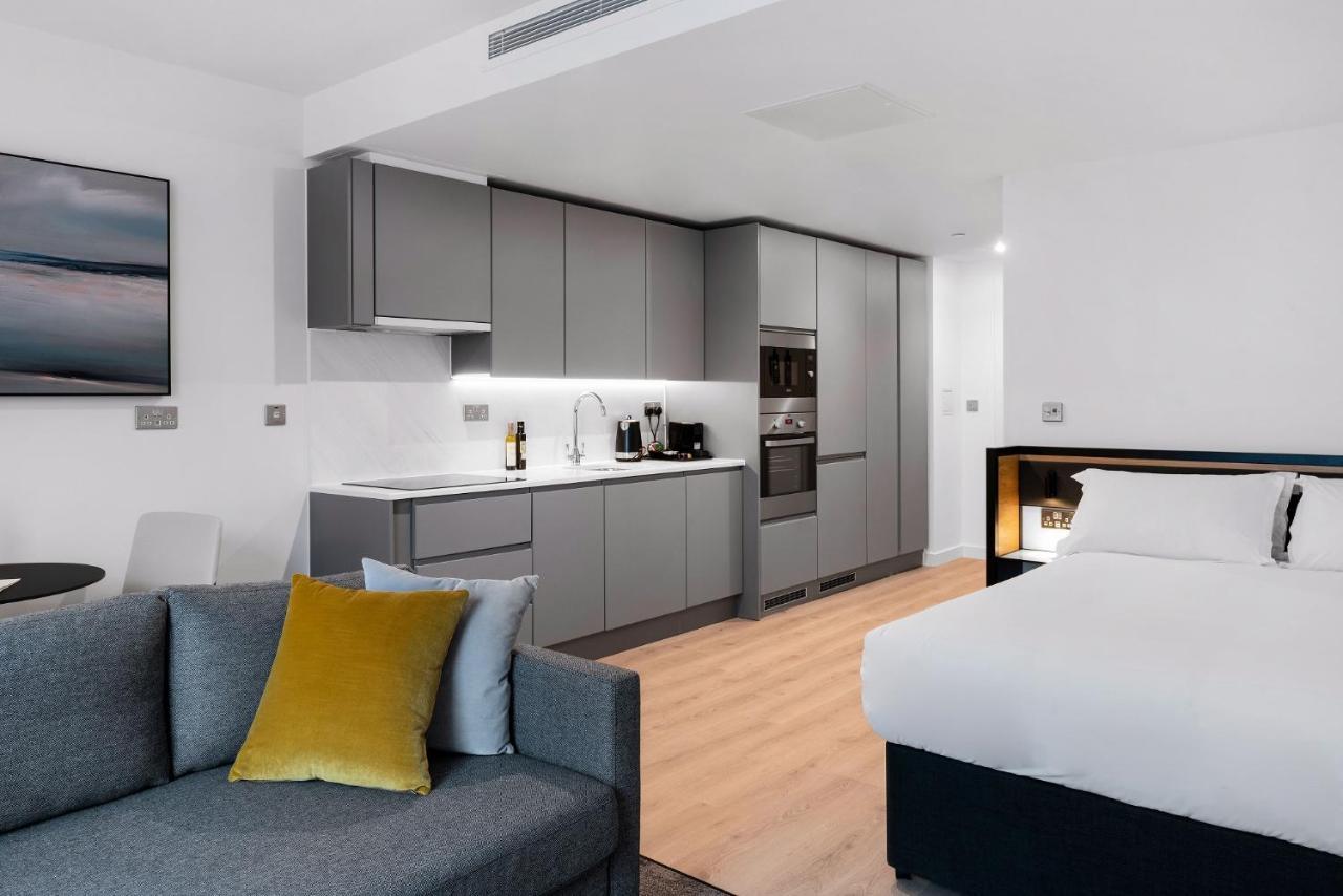 Citysuites 2 Aparthotel Manchester Kültér fotó The photo shows a modern studio apartment featuring an open-plan layout. On one side, there is a kitchenette with sleek, gray cabinets and appliances, including a coffee maker and oven. Adjacent to the kitchenette is a small dining area with a table 