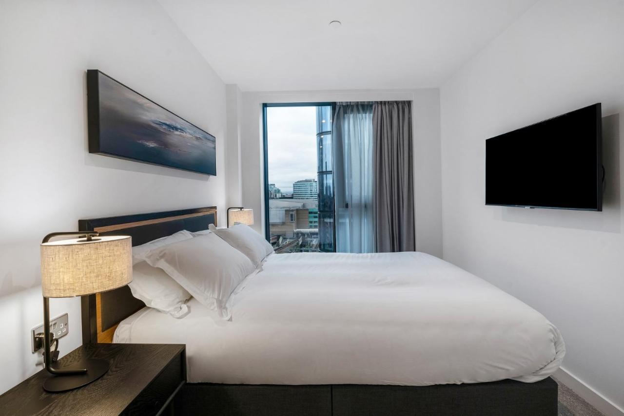 Citysuites 2 Aparthotel Manchester Kültér fotó The photo shows a modern bedroom with a minimalist aesthetic. It features a large bed with white bedding and two pillows. On either side of the bed, there are bedside tables with lamps. A large window lets in natural light, covered by sheer curtains.
