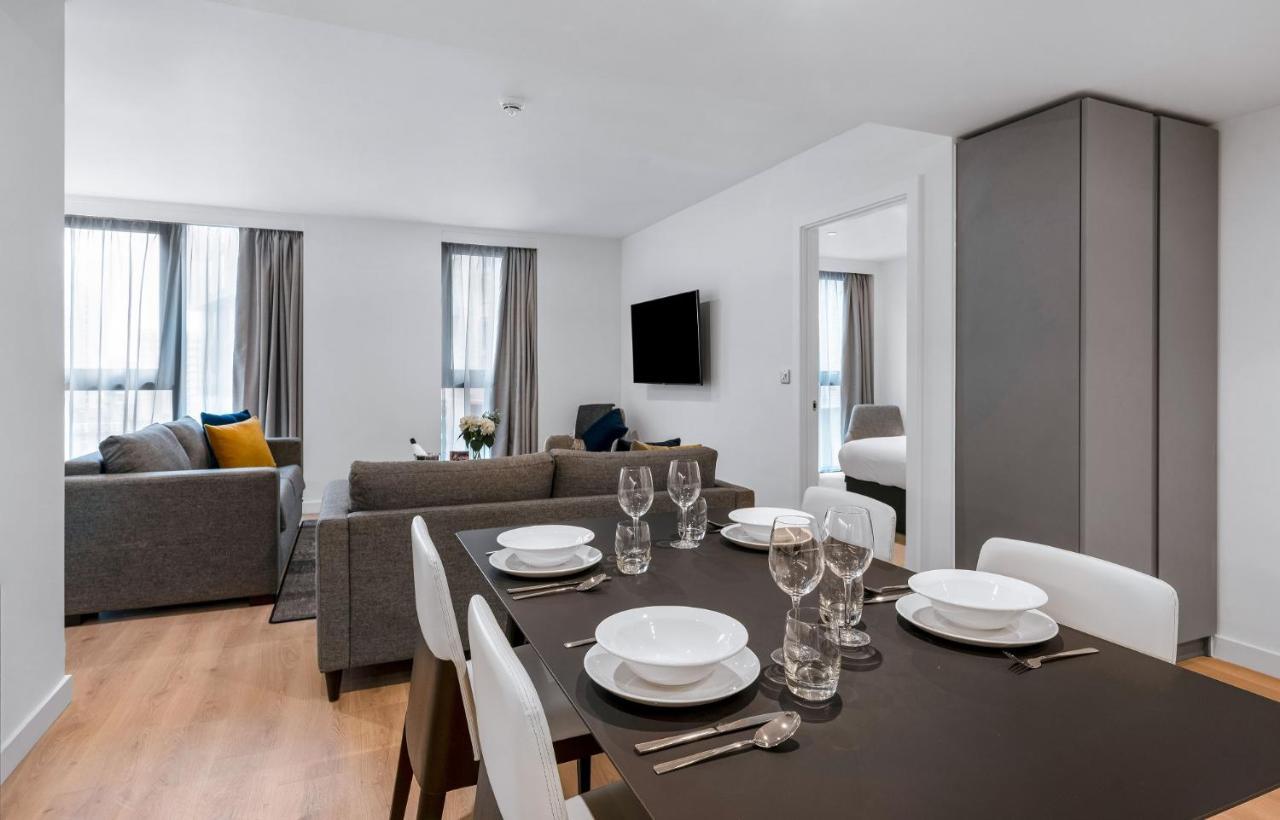 Citysuites 2 Aparthotel Manchester Kültér fotó The photo shows a modern living space with an open layout. In the foreground, there is a dining table set for a meal, featuring multiple plates, glasses, and cutlery arranged neatly. In the background, there is a comfortable-looking gray sofa with a 