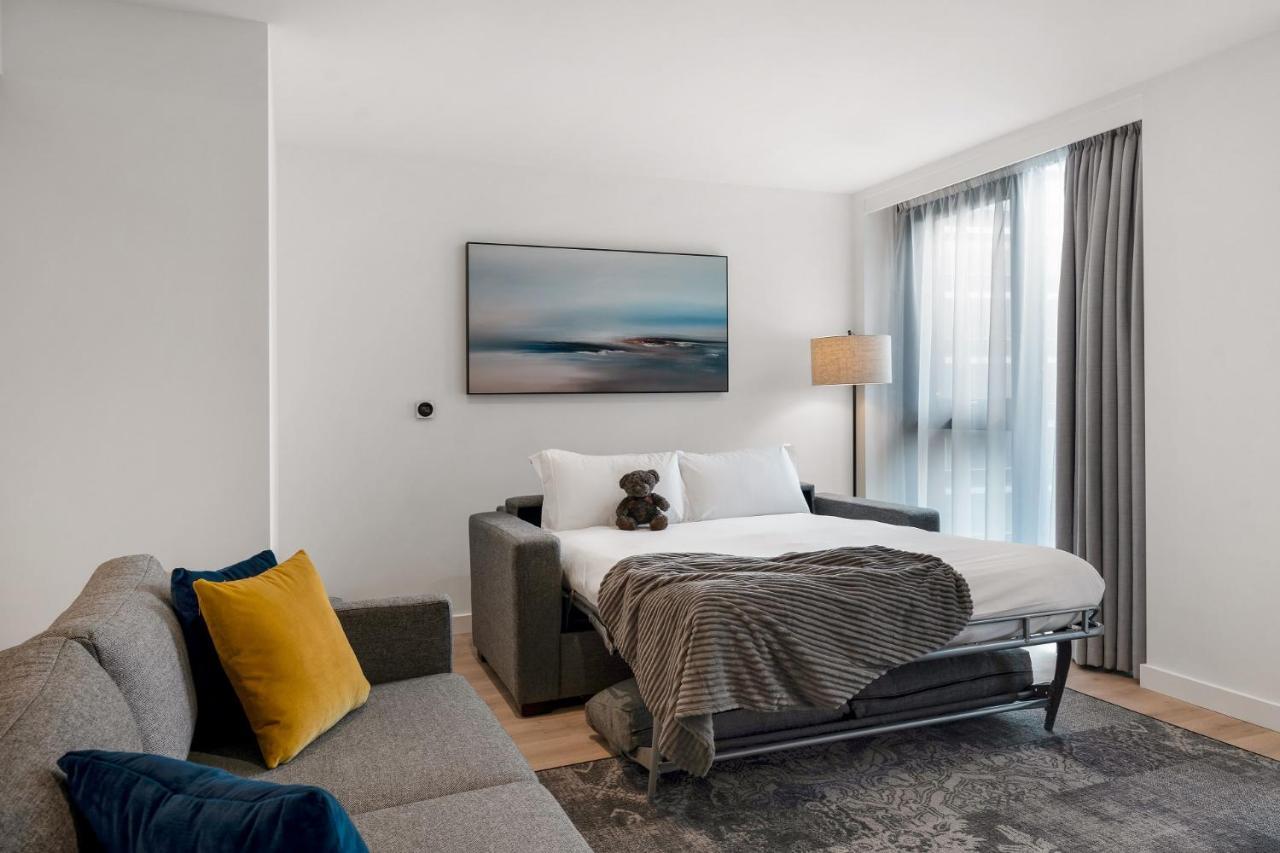 Citysuites 2 Aparthotel Manchester Kültér fotó The photo shows a modern and minimalistic guest room. There is a sofa on the left side, adorned with a colorful cushion. In the middle is a bed that appears to be a pull-out style, featuring white bedding and a gray throw blanket. A stuffed toy is pl
