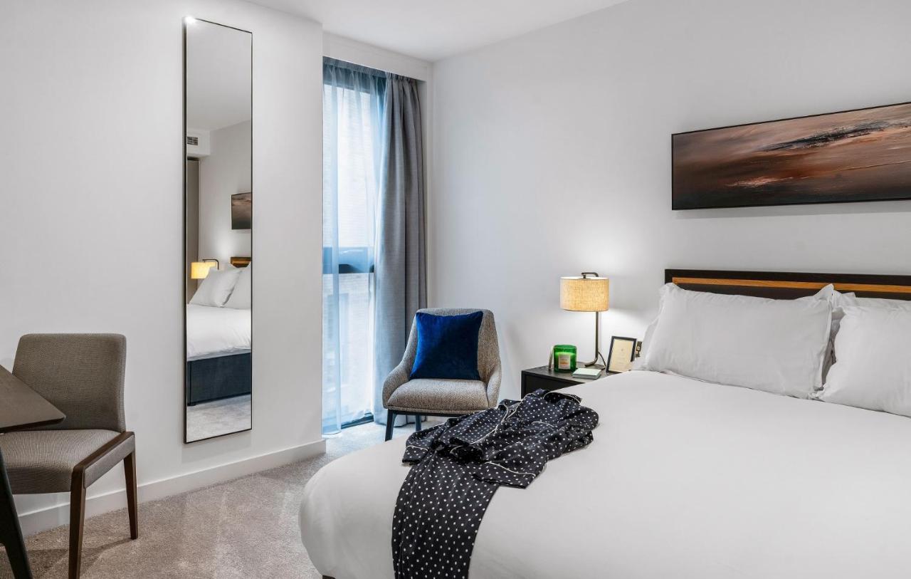 Citysuites 2 Aparthotel Manchester Kültér fotó The photo shows a modern hotel room. It features a neatly made bed with white linens and a dark-colored throw draped over it. There is a small bedside table with a lamp next to the bed. A comfortable chair in a light color is positioned near the wind