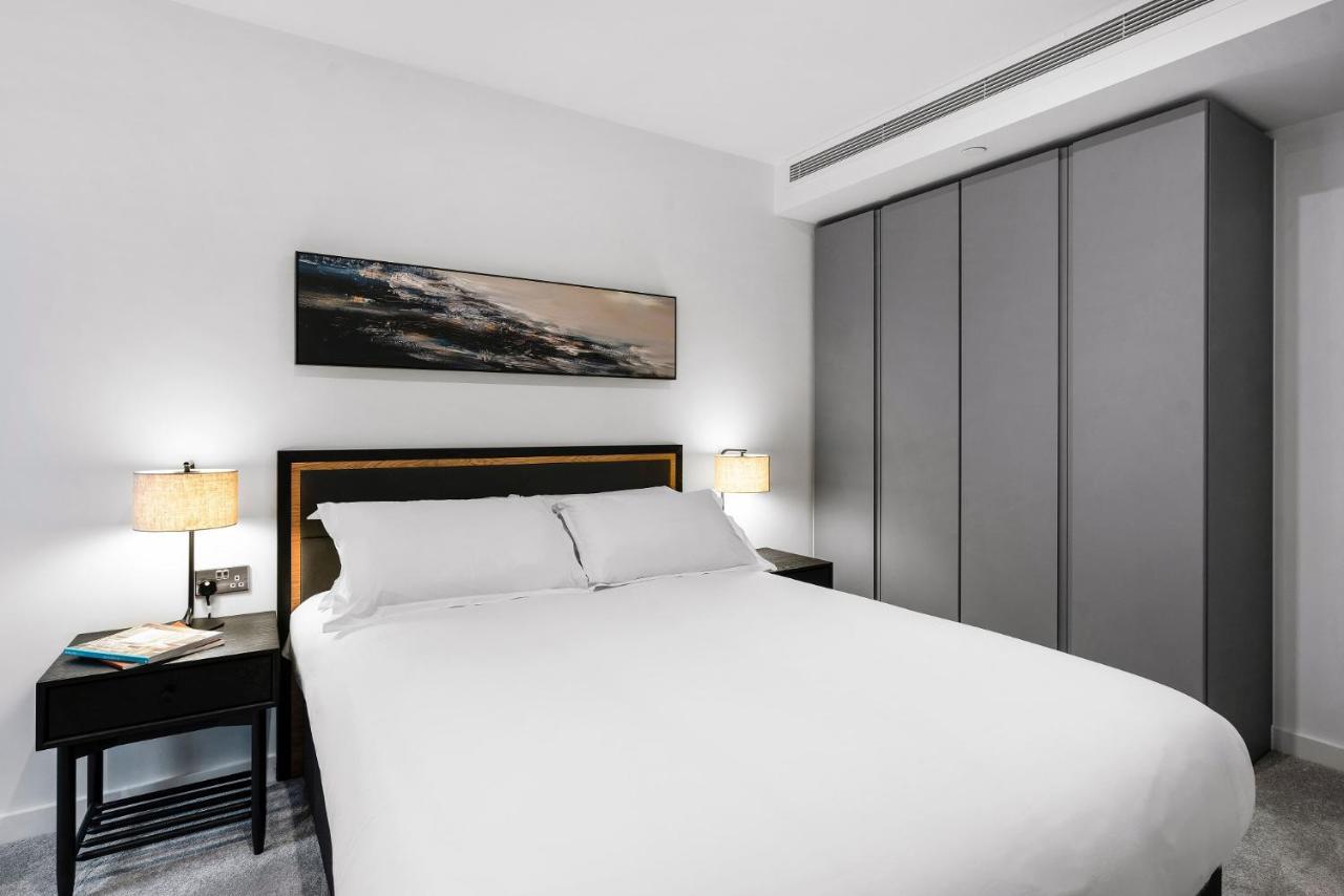 Citysuites 2 Aparthotel Manchester Kültér fotó The photo shows a modern bedroom designed with a minimalist aesthetic. The focal point is a large bed with white bedding, positioned against a wall. On either side of the bed, there are black nightstands topped with lamps, providing a warm glow. Abov