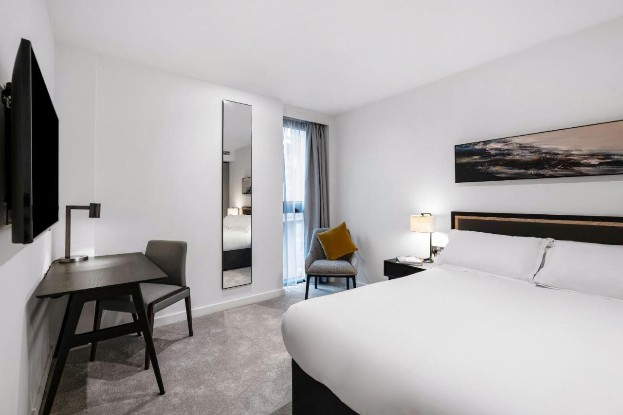 Citysuites 2 Aparthotel Manchester Kültér fotó The photo shows a modern hotel room with a clean and minimalist design. It features a large bed with white linens, a small desk with a chair, and a mirror mounted on the wall. There is a chair in one corner with a decorative pillow, and the room has 