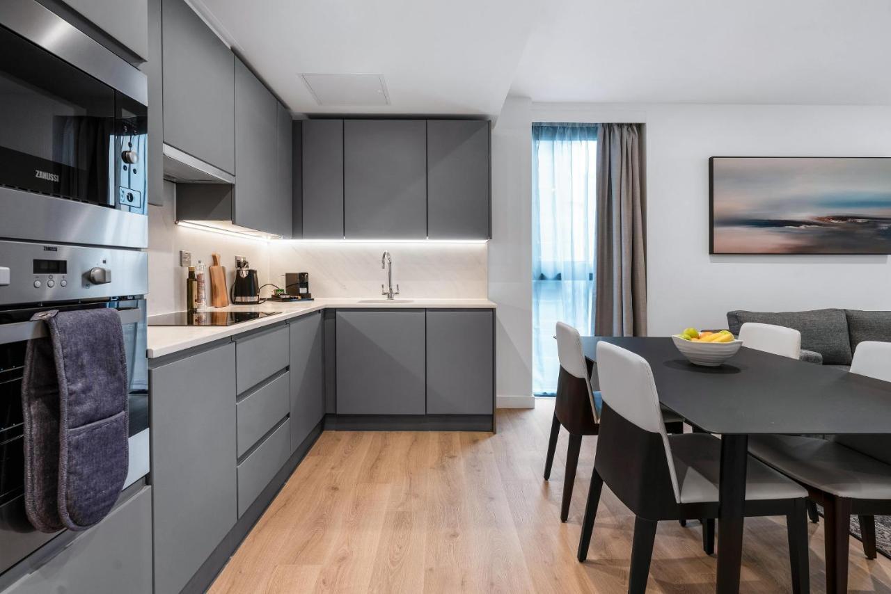 Citysuites 2 Aparthotel Manchester Kültér fotó The photo shows a modern kitchen and dining area. The kitchen features sleek gray cabinetry, a white countertop, and built-in appliances such as an oven. There is a sink and some kitchen utensils displayed. The adjacent dining area contains a black t