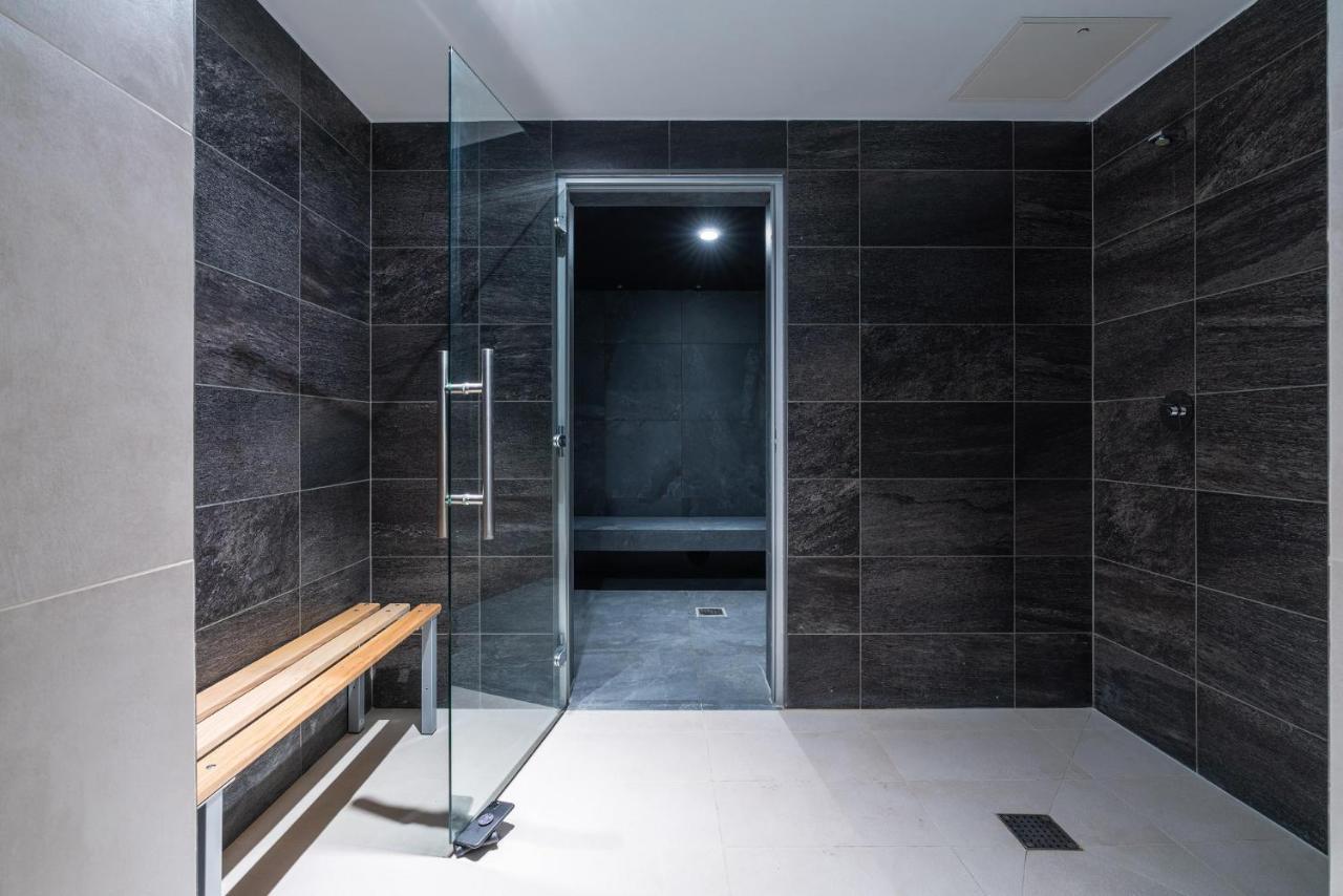 Citysuites 2 Aparthotel Manchester Kültér fotó The photo shows a modern, minimalist shower area. The space is predominantly tiled with dark gray tiles on the walls, giving it a sleek and sophisticated look. There is a glass door that leads to another room, and a wooden bench is positioned against