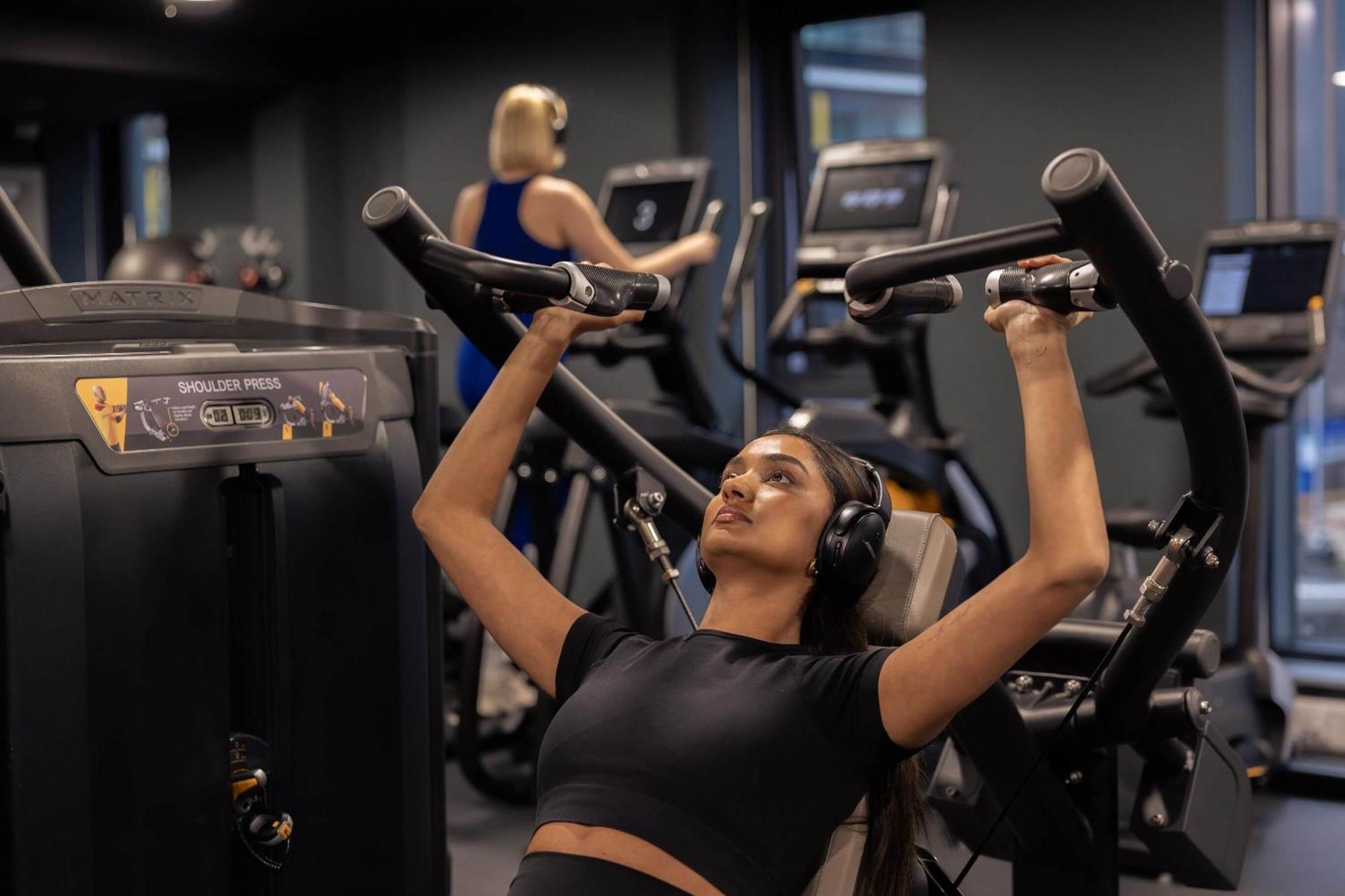 Citysuites 2 Aparthotel Manchester Kültér fotó The photo shows a gym setting with two individuals engaged in physical exercise. In the foreground, a woman is using a chest press machine while wearing headphones, focusing on her workout. She appears to be in a seated position, pushing the handles 