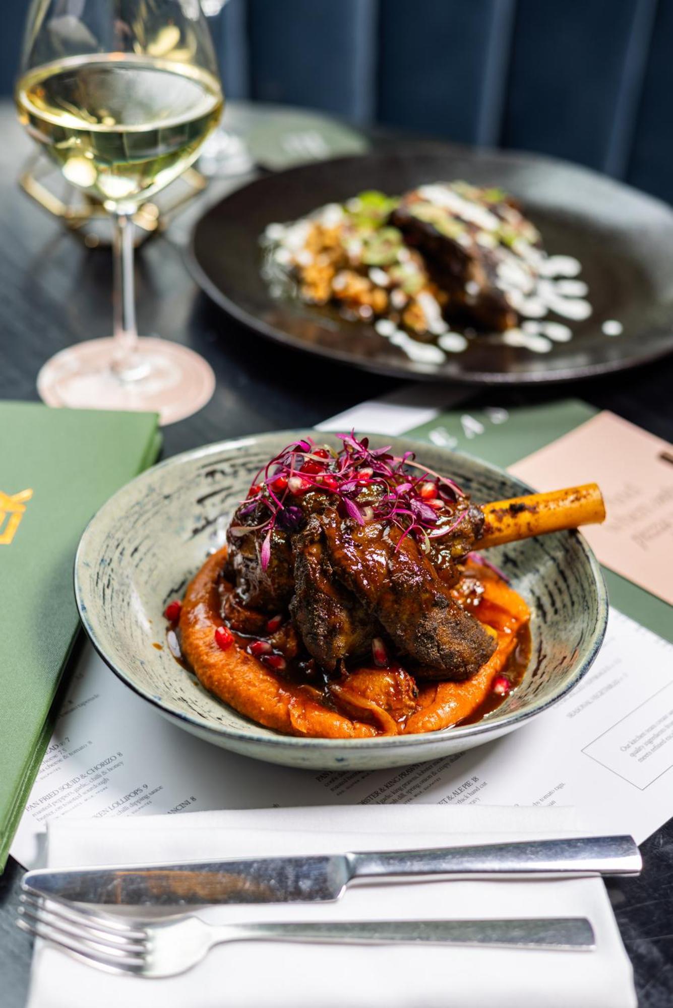 Citysuites 2 Aparthotel Manchester Kültér fotó The photo showcases a beautifully presented dish featuring a piece of slow-cooked meat with a bone, likely lamb, sitting atop a vibrant orange puree. The meat is glazed with a rich sauce and garnished with small pieces of red pomegranate and purple m