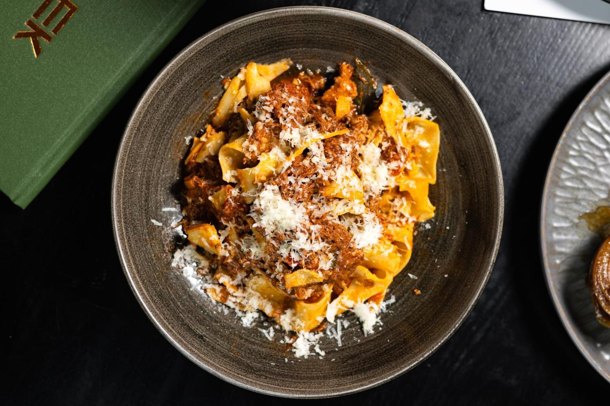 Citysuites 2 Aparthotel Manchester Kültér fotó The photo shows a bowl of pasta featuring wide, ribbon-like noodles, likely pappardelle, fully coated in a rich meat sauce. The dish is garnished with a generous sprinkling of grated cheese, adding a creamy texture to the presentation. The dark bowl 