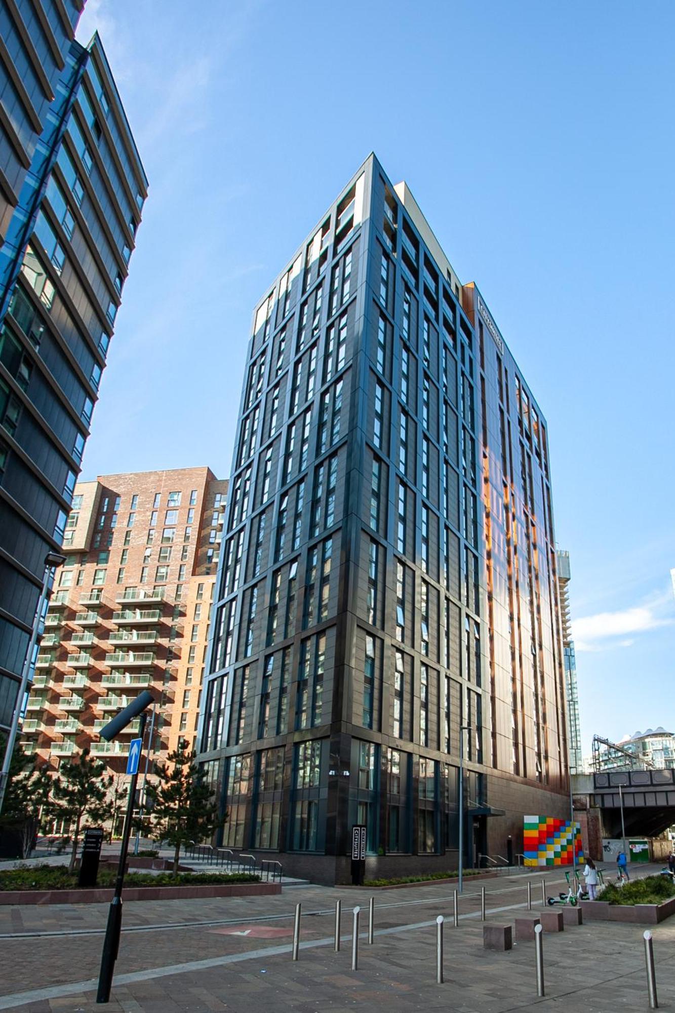 Citysuites 2 Aparthotel Manchester Kültér fotó The photo shows a modern high-rise building with a sleek, glass façade. It is set against a blue sky, indicating a clear day. The building appears to have multiple floors with large windows that reflect light. Nearby, there are additional buildings, 